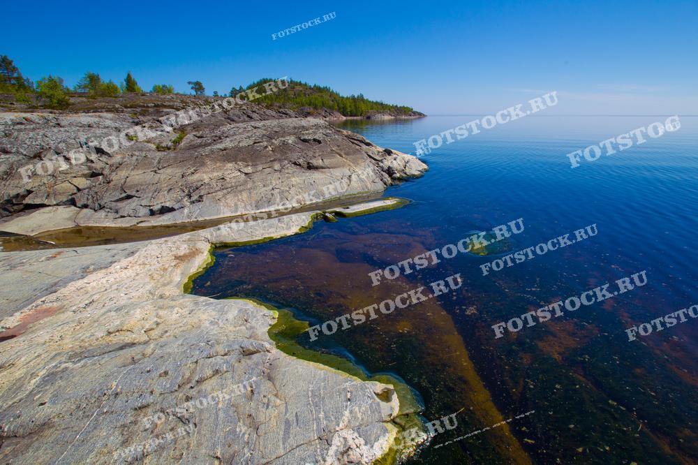 На фотографии изображён скалистый берег Ладожского озера в Карелии. Прозрачные голубые воды омывают каменистые поверхности, создавая живописный пейзаж. Ясный летний день и безоблачное небо подчеркивают природную красоту этого региона. Ладожское озеро славится своими чистыми водами и уникальными скалистыми берегами, привлекая множество туристов, желающих насладиться уединением и природной гармонией.